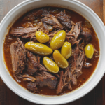 Mississippi pot roast freshly cooked and served in a white ceramic dish, featuring tender shredded beef in rich gravy with pepperoncini peppers and butter, placed on a rustic wooden table.